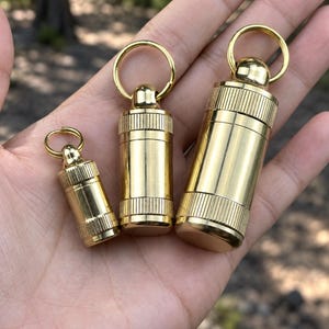 Könnte beinhalten: Drei zylindrische Pillenbehälter aus Messing in verschiedenen Größen, jeweils mit einem Schraubdeckel mit geriffeltem Griff und einem kreisförmigen Metallring an der Oberseite für Schlüsselanhänger. Die polierten goldfarbenen Metalloberflächen sind glatt.