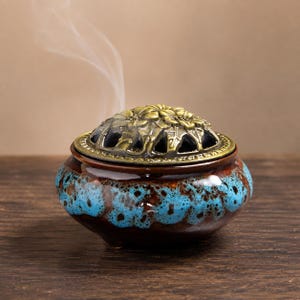 May include: A round ceramic incense burner with a glossy brown and bright blue drip glaze finish. The vessel features a removable, ornate brass-toned metal lid with a cut-out floral pattern, from which a thin wisp of white smoke rises upward.