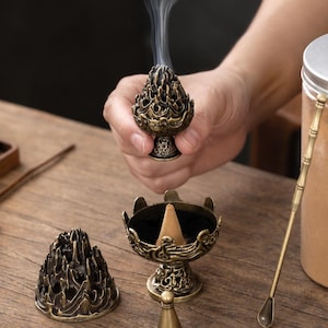 Puede incluir: Un set de quemador de incienso de metal en tono bronce que incluye una tapa decorativa calada con textura de montaña, una base de cuenco a juego con un cono de incienso color canela, una pequeña herramienta cónica y una cuchara de metal larga.