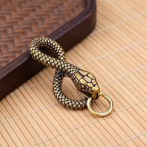 May include: A brass-toned metal keyring shaped like a coiled snake with a textured, scale-like surface pattern. The snake's head is positioned at the front, and a smooth, circular metal ring is attached to the lower jaw of the snake figure.