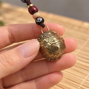 Op de afbeelding: Een koperen schildpadschild-bedel hangend aan een donkerbruin gevlochten koord. Het koord is voorzien van een reeks decoratieve kralen, waaronder een zwarte kraal met een wit wolkenmotief, een gespikkelde beige kraal en een donkerrode kraal.