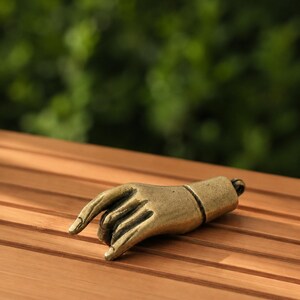 May include: A brass-colored metal pendant shaped like a hand with fingers curled into a specific gesture. The object has a smooth, metallic finish and features a small circular loop at the base, allowing it to be attached to a chain or cord.