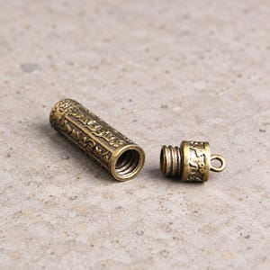 May include: A cylindrical brass pendant container with a screw-top lid featuring an attached loop. The exterior surface of both the main body and the cap is engraved with intricate script characters in a dark, weathered metallic finish.