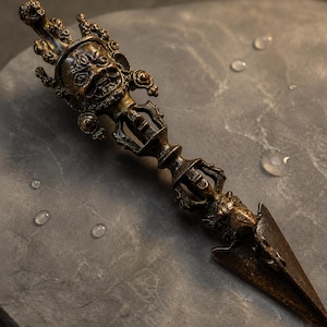 May include: A bronze-toned phurba ritual dagger featuring an intricate, multi-faced deity head at the top, a central handle with ornate openwork detailing, and a sharp, triangular blade tip extending from a stylised animal head base.