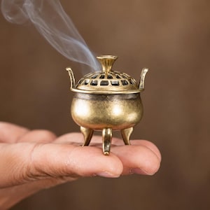 Puede incluir: Un pequeño quemador de incienso de metal color latón, de forma redonda, con tres patas curvas, dos asas laterales y una tapa extraíble con un patrón de rejilla recortado. Penachos de humo blanco se elevan desde la parte superior.