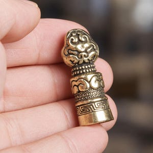 May include: A small, cylindrical brass object featuring intricate relief carvings. The top section displays a stylized cloud motif, followed by a band of beaded texture, a middle section with swirling patterns, and a base with a geometric Greek key design.