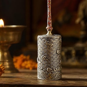 Puede incluir: Un molinillo de oración cilíndrico de metal con un intrincado patrón en relieve de motivos de olas repetitivas y corrientes de agua. El objeto está suspendido por un cordón trenzado multicolor unido al centro superior del cuerpo plateado.