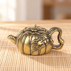 May include: A small, metallic gold-toned teapot shaped like a pumpkin. The body features vertical ridges, a central stem-like lid handle, a curved spout, and a decorative handle with an embossed leaf design on the side of the vessel.