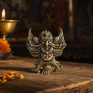 Op de afbeelding: Een messingkleurig metalen beeldje van Garuda, een mythisch vogelwezen, met ingewikkelde vleugelpatronen, een scherpe snavel en gedetailleerde schubstructuren op het onderlichaam, staand in een symmetrische houding met de armen gespreid.