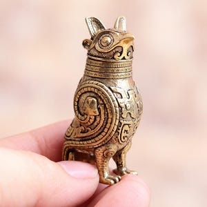 May include: A small, metallic, gold-toned figurine shaped like an owl with intricate, embossed patterns covering its entire surface. The owl features large, circular eyes, pointed ears, and detailed scrollwork designs on its wings and chest.