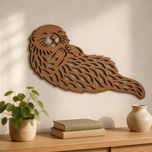 Op de afbeelding: Een bruin houten wanddecoratie in de vorm van een zeeotter die op zijn rug drijft. Het uitgesneden ontwerp bevat ingewikkelde, vloeiende lijnen die de vachtstructuur, gelaatstrekken en pootjes van de otter definiëren voor een gedetailleerd silhouet.