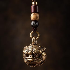 May include: A brass pendant shaped like a stylized creature head with intricate engravings, suspended from a brown cord. The cord features a gold-toned metal bead, a dark red wooden bead, a speckled beige bead, and a black bead with a white cloud icon.
