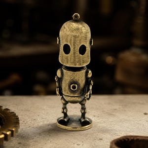 May include: A small, standing robot figurine crafted from a metallic material with a weathered, antique brass finish. The robot features a rounded head with two dark, oval-shaped eye cutouts, a cylindrical torso with a central circular detail, and thin limbs.