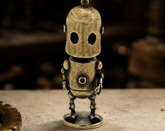 May include: A small, standing robot figurine crafted from a metallic material with a weathered, antique brass finish. The robot features a rounded head with two dark, oval-shaped eye cutouts, a cylindrical torso with a central circular detail, and thin limbs.