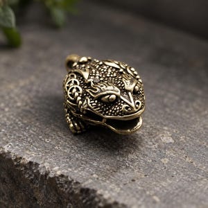 May include: A brass-colored metal pendant shaped like a toad with a textured, bumpy surface and intricate engraved details on its head and back. The toad has a wide, open mouth and a small loop at the top for attachment, featuring a metallic finish.