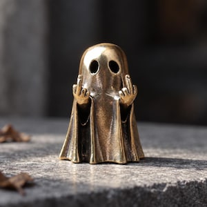 Könnte beinhalten: Eine kleine, metallische, messingfarbene Geisterfigur. Der Geist hat zwei hohle, ovale Augen und ist mit erhobenen Armen dargestellt, wobei er an beiden Händen den Mittelfinger zeigt. Die Oberfläche hat ein verwittertes, antikes Finish.