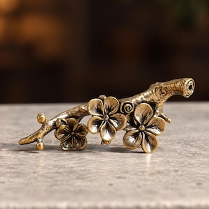 May include: A metallic gold-toned decorative object shaped like a tree branch with three detailed five-petaled flowers and small buds. The surface has a textured, antique finish with dark recessed areas highlighting the floral and branch details.