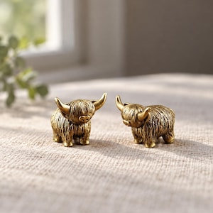May include: Two small, gold-toned metal figurines shaped like Highland cattle. Each figurine features a textured, shaggy coat, curved horns, and a detailed face with a prominent snout. The figures are standing side by side, facing slightly inward.