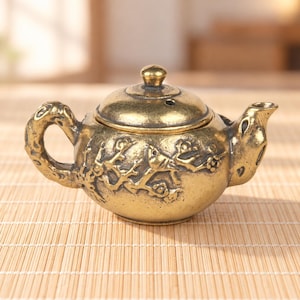 May include: A small, antique-style brass teapot featuring a textured, relief-carved surface with floral branch motifs. The teapot has a rounded body, a curved handle, a short spout, and a removable lid with a small knob and a steam vent hole.