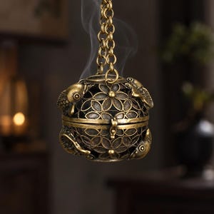 May include: A spherical brass incense burner featuring an intricate openwork floral pattern and three sculpted fish figures attached to the sides. The metal sphere is suspended by a matching brass chain, with thin wisps of smoke rising from the top.