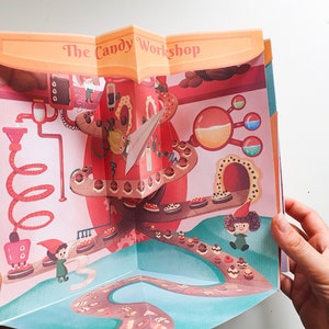 May include: A pop-up book with a colorful illustration of a candy factory. The book is open to reveal a 3D scene with a candy conveyor belt, a red and white candy machine, and a small gnome figure. The title of the book is "The Candy Workshop."
