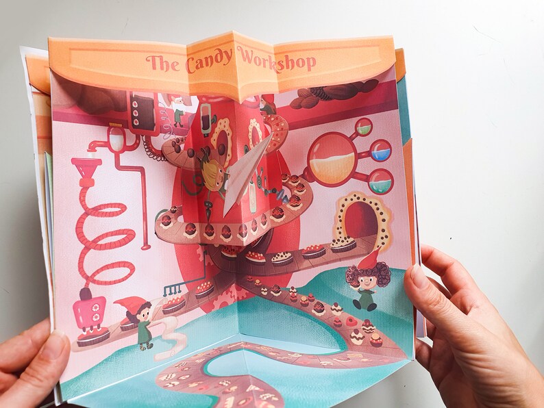 May include: A pop-up book with a colorful illustration of a candy factory. The book is open to reveal a 3D scene with a candy conveyor belt, a red and white candy machine, and a small gnome figure. The title of the book is "The Candy Workshop."