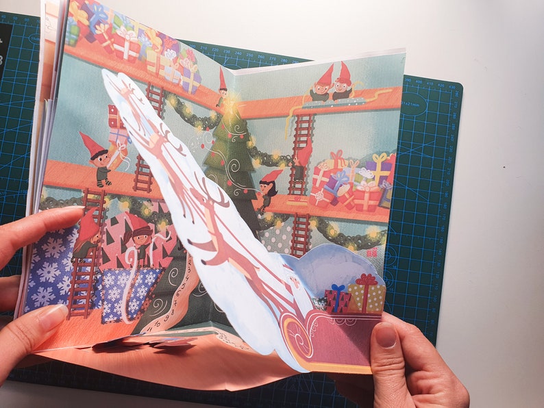 May include: A pop-up book with a Christmas theme. The book features a reindeer pulling a sleigh full of presents. The reindeer is made of paper and is attached to the book with a string. The sleigh is also made of paper and is filled with colorful presents. The book is open to a page that shows a group of elves working on a Christmas tree.