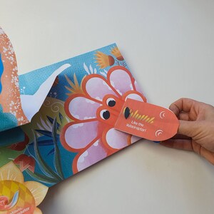 May include: A colorful pop-up book with a dinosaur theme. The open page features a large, orange dinosaur head with the text "Like the Velociraptor!" and a detailed floral background. A hand is holding the dinosaur's head.