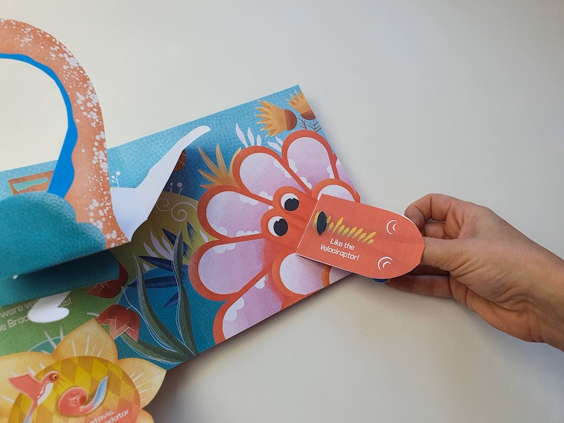 May include: A colorful pop-up book with a dinosaur theme. The open page features a large, orange dinosaur head with the text "Like the Velociraptor!" and a detailed floral background. A hand is holding the dinosaur's head.