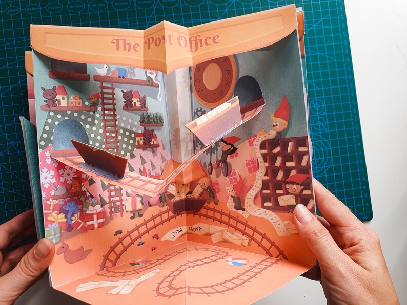 May include: A pop-up book with a festive Christmas theme. The book is open to a scene of a post office with elves working and a train track with a toy train. The text on the book reads "The Post Office".