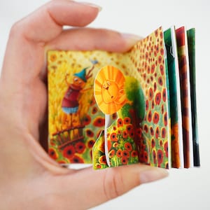 Può includere: Un piccolo libro pop-up aperto con illustrazioni colorate. Le pagine raffigurano una scena stravagante con un personaggio dei cartoni animati, girasoli e papaveri. Il libro è tenuto aperto, mostrando le vivaci opere d'arte.
