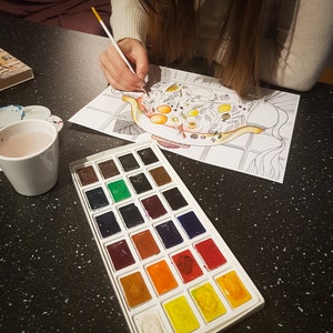 May include: A watercolor paint set with 24 colors in a white plastic tray. The tray is on a black surface with a coloring book page showing a teapot with flowers and fruit.