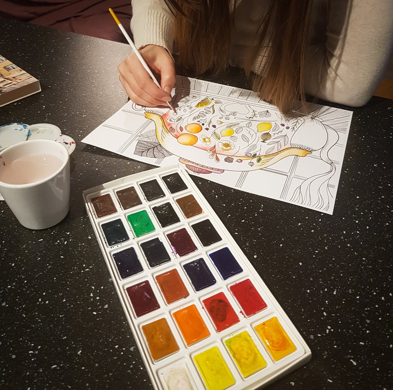 May include: A watercolor paint set with 24 colors in a white plastic tray. The tray is on a black surface with a coloring book page showing a teapot with flowers and fruit.