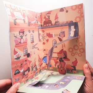 May include: A pop-up book with a colorful illustration of a workshop filled with elves making toys. The book is open to reveal a 3D pop-up of an elf playing the cello.