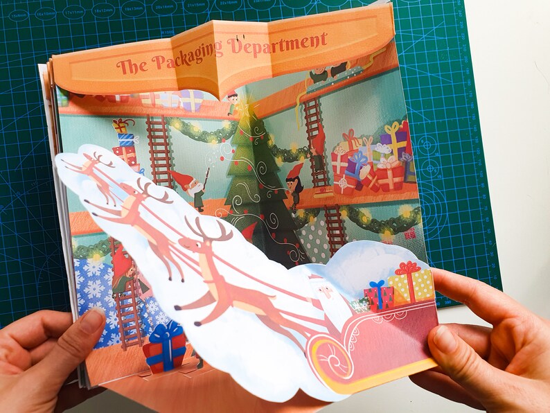 May include: A pop-up book with a festive Christmas scene. The book features a red and white sleigh pulled by reindeer, with a white figure of Santa Claus in the sleigh. The book's cover reads "The Packaging Department".