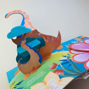 May include: A colorful pop-up book featuring a dinosaur theme. The open pages display a 3D Brachiosaurus with a long neck and a blue cloud. Text on the page reads "Some were very tall! Like the Brachiosaurus!"