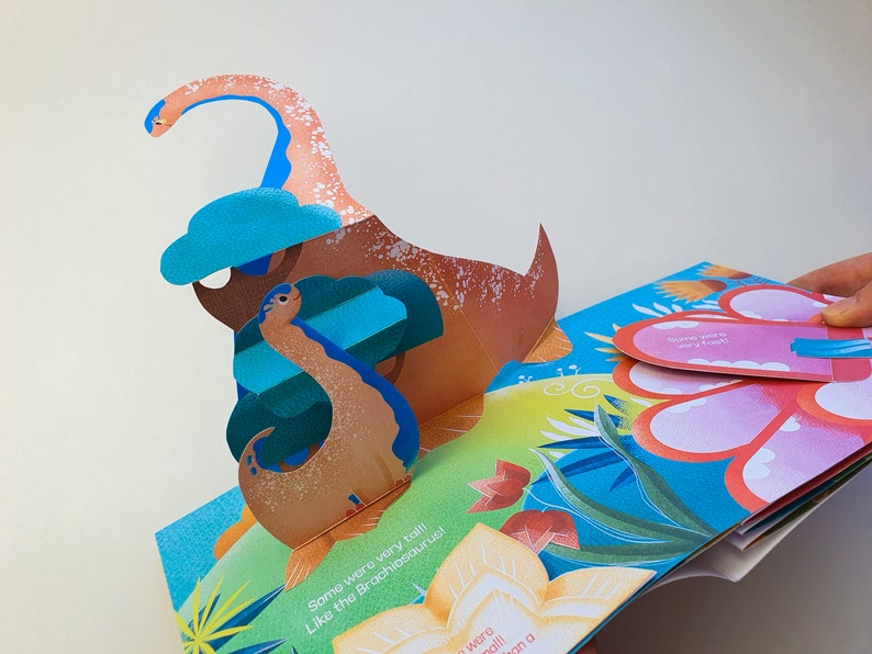 May include: A colorful pop-up book featuring a dinosaur theme. The open pages display a 3D Brachiosaurus with a long neck and a blue cloud. Text on the page reads "Some were very tall! Like the Brachiosaurus!"