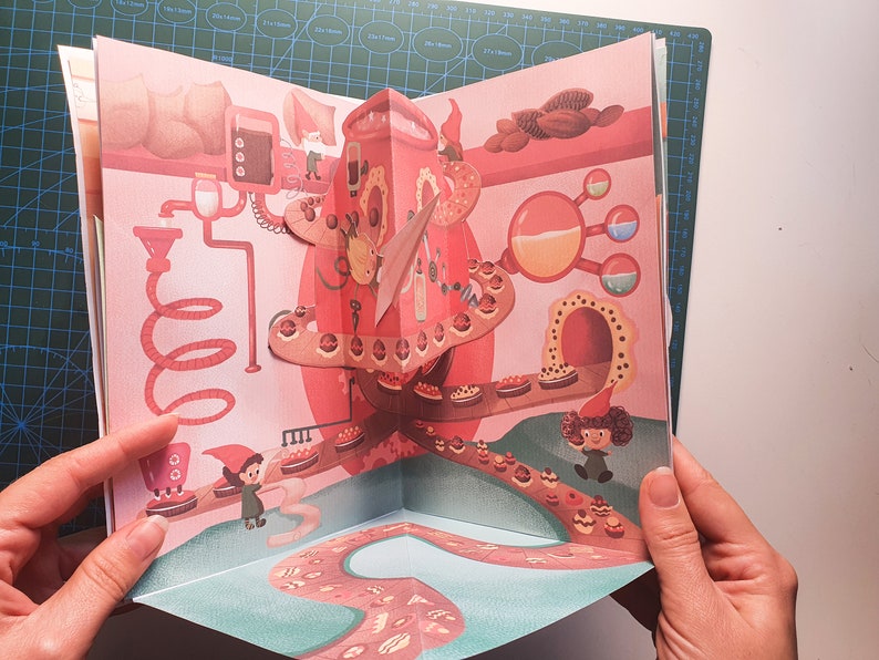 May include: A pop-up book with a 3D illustration of a chocolate factory. The factory is made of red and pink cardboard and features a conveyor belt with chocolate pastries. There are two small gnome figures in the scene.