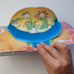 May include: A pop-up children's book with a dinosaur theme. The open page reveals two green dinosaurs in a landscape with a blue river. The text "Guess what? All dinosaurs came from eggs!" is visible.