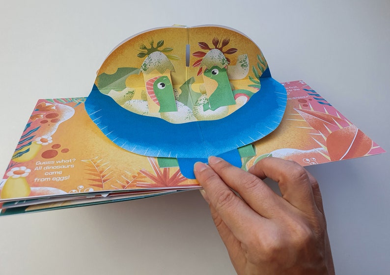 May include: A pop-up children's book with a dinosaur theme. The open page reveals two green dinosaurs in a landscape with a blue river. The text "Guess what? All dinosaurs came from eggs!" is visible.