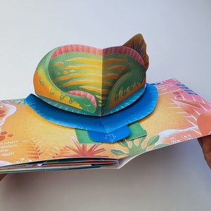 May include: A colorful pop-up book with a dinosaur theme. The open pages reveal a 3D dinosaur figure in shades of green, orange, and yellow. Text on the page reads, "Guess what? All dinosaurs come from eggs!"