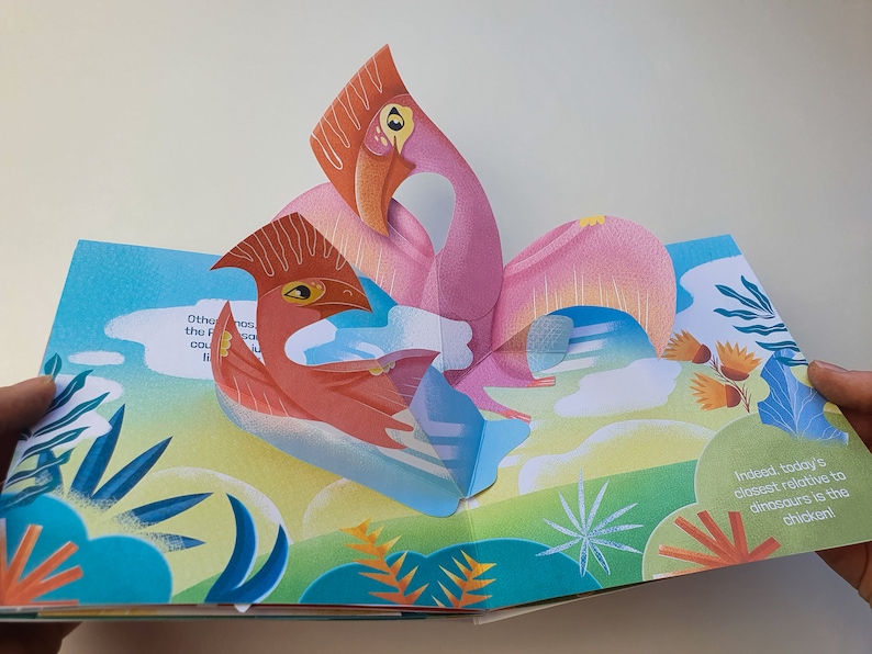 May include: A pop-up book featuring two stylized pterodactyls in shades of pink and red against a backdrop of a blue sky and green landscape. The book is open, revealing the 3D paper art and text about dinosaurs and chickens.