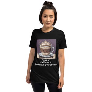 Op de afbeelding: Zwart T-shirt met een cartoonillustratie van een kopje koffie met slagroom en de tekst "Runs on Caffeine & Executive Dysfunction".