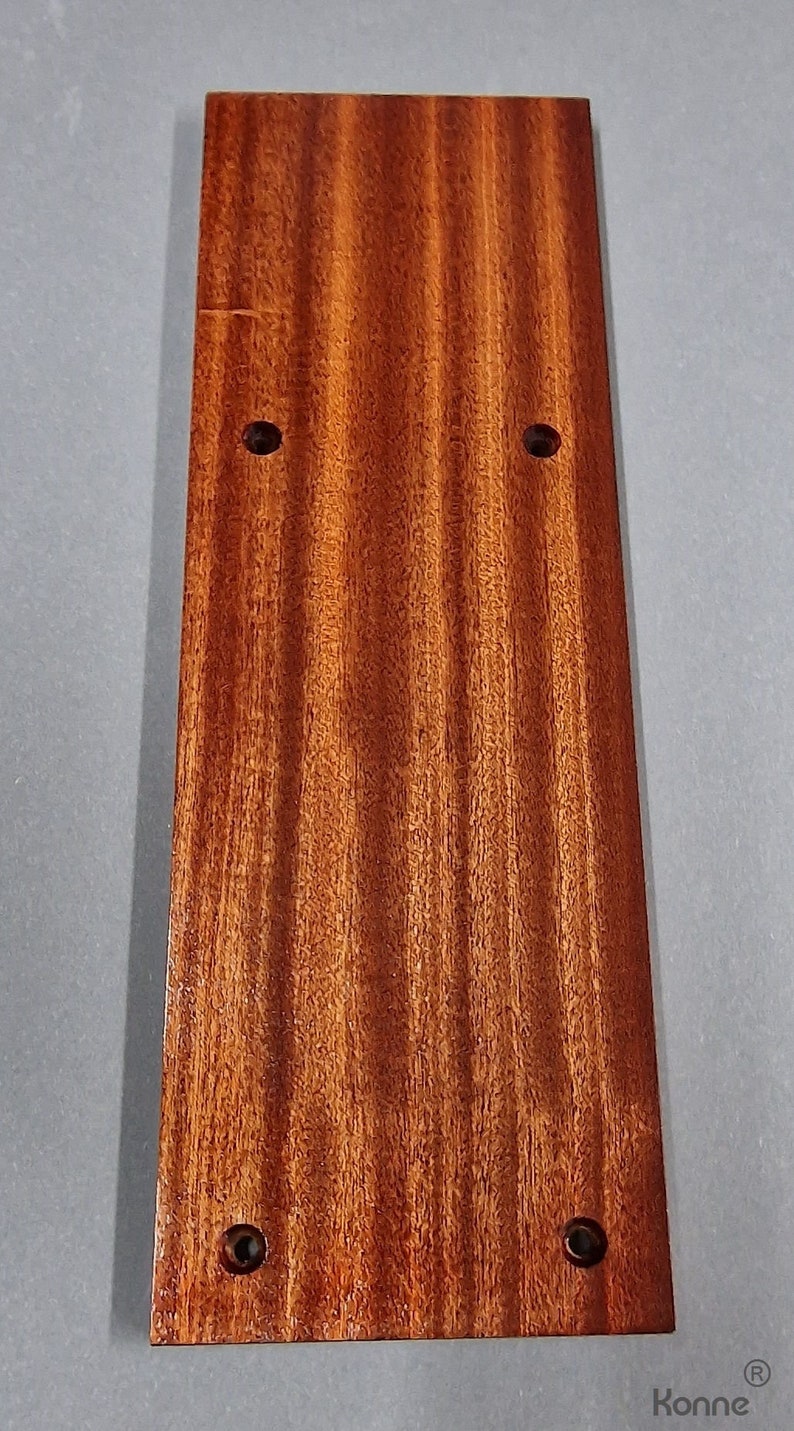 Puede incluir: Una placa rectangular de madera de color marr&oacute;n rojizo intenso con un patr&oacute;n de veta de madera. La placa tiene cuatro agujeros perforados a intervalos regulares.