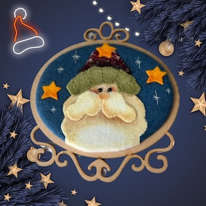 May include: A felt Christmas ornament featuring Santa Claus with a cream-colored beard and a green and patterned hat. The ornament is set against a blue background with gold stars and a decorative frame. A festive holiday decoration.
