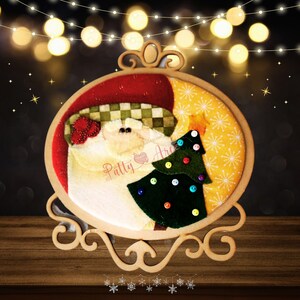 May include: A wooden Christmas ornament featuring a Santa Claus design with a green Christmas tree. The ornament has a wooden frame with a decorative base and top. The Santa Claus has a red hat and a checkered pattern.