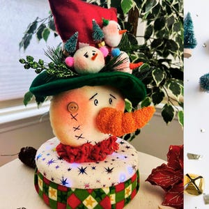 May include: A whimsical snowman decoration with a red felt hat and a green brimmed hat. The snowman has an orange carrot nose, button eyes, and a stitched mouth. It is adorned with miniature snowmen and Christmas trees.
