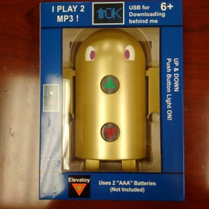 May include: A gold-coloured toy elevator with a green up arrow and a red down arrow. The toy has a digital display that reads "OK" and says "I Play 2 MP3!" on the box. The box also says "Uses 2 "AAA" Batteries (Not Included)" and "USB for Downloading behind me".