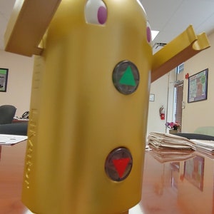 May include: A gold robot toy with a green triangle and red triangle on its body. The robot has two arms and two legs. The robot is standing on a wooden surface.