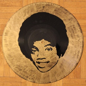 May include: A gold vinyl record with a black and white silhouette of a person's face. The person has short, curly hair and is looking directly at the viewer.